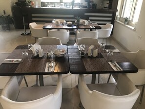 A dining area with tables set for two, white chairs, and a kitchen in the background.