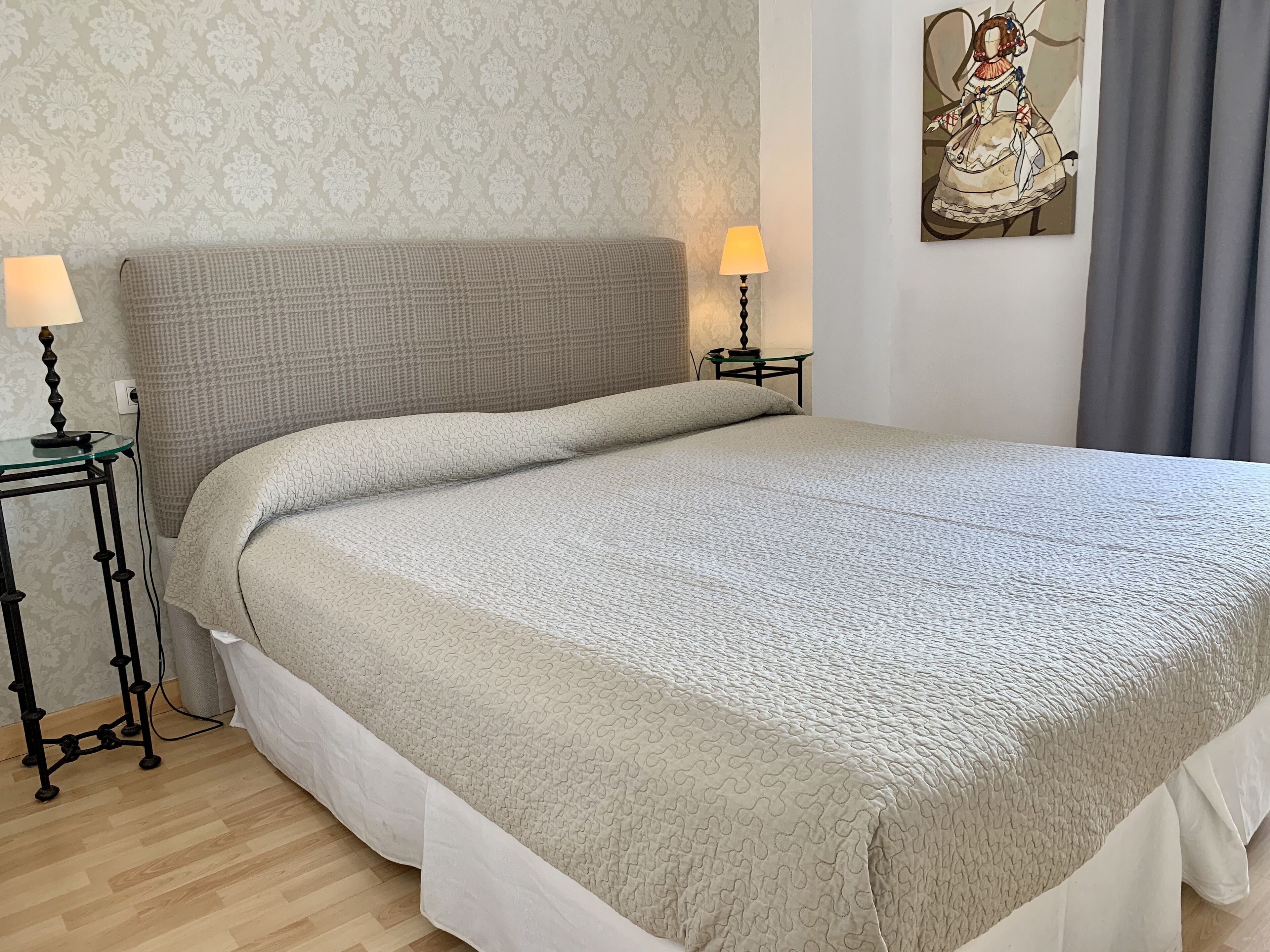 A neatly made bed with a textured blanket, two bedside lamps, a small table with a book, and a wall painting.