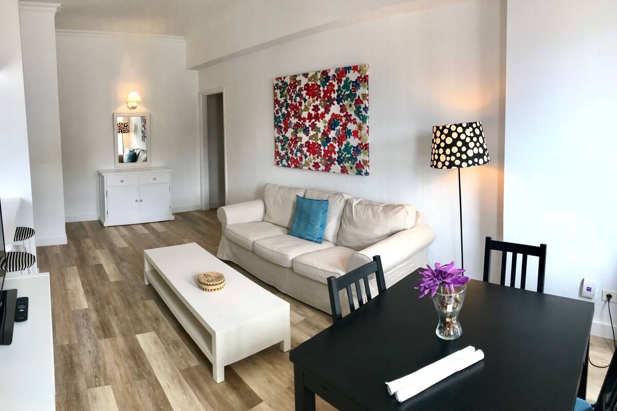 Ein modernes Wohnzimmer mit einem weißen Sofa, einem Couchtisch, einem schwarzen Esstisch mit Blumenvase und einem farbenfrohen Blumenbild an der Wand.