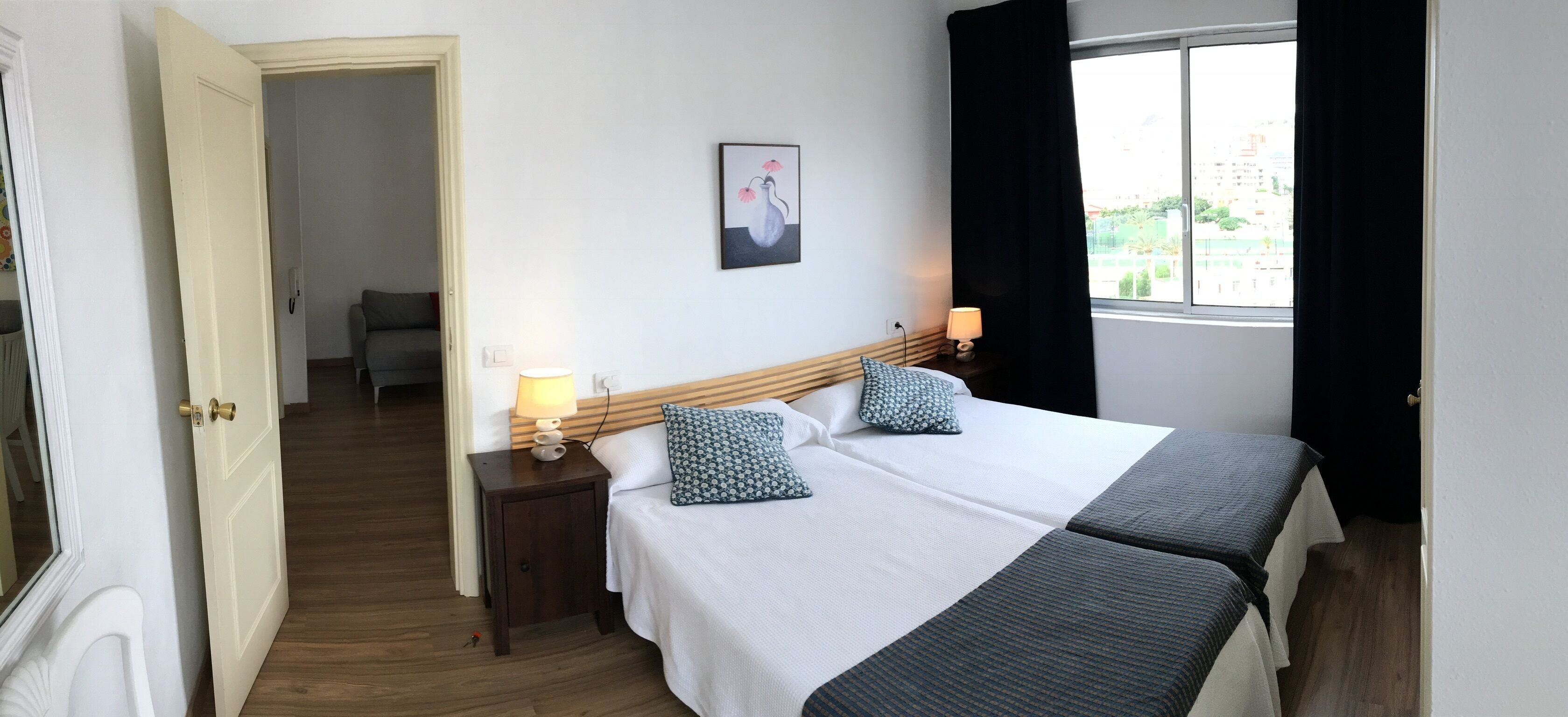 A hotel room with a double bed, a bedside table with a lamp, a painting on the wall, and a window with curtains.
