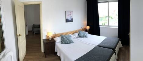 A hotel room with a double bed, a bedside table with a lamp, a painting on the wall, and a window with curtains.