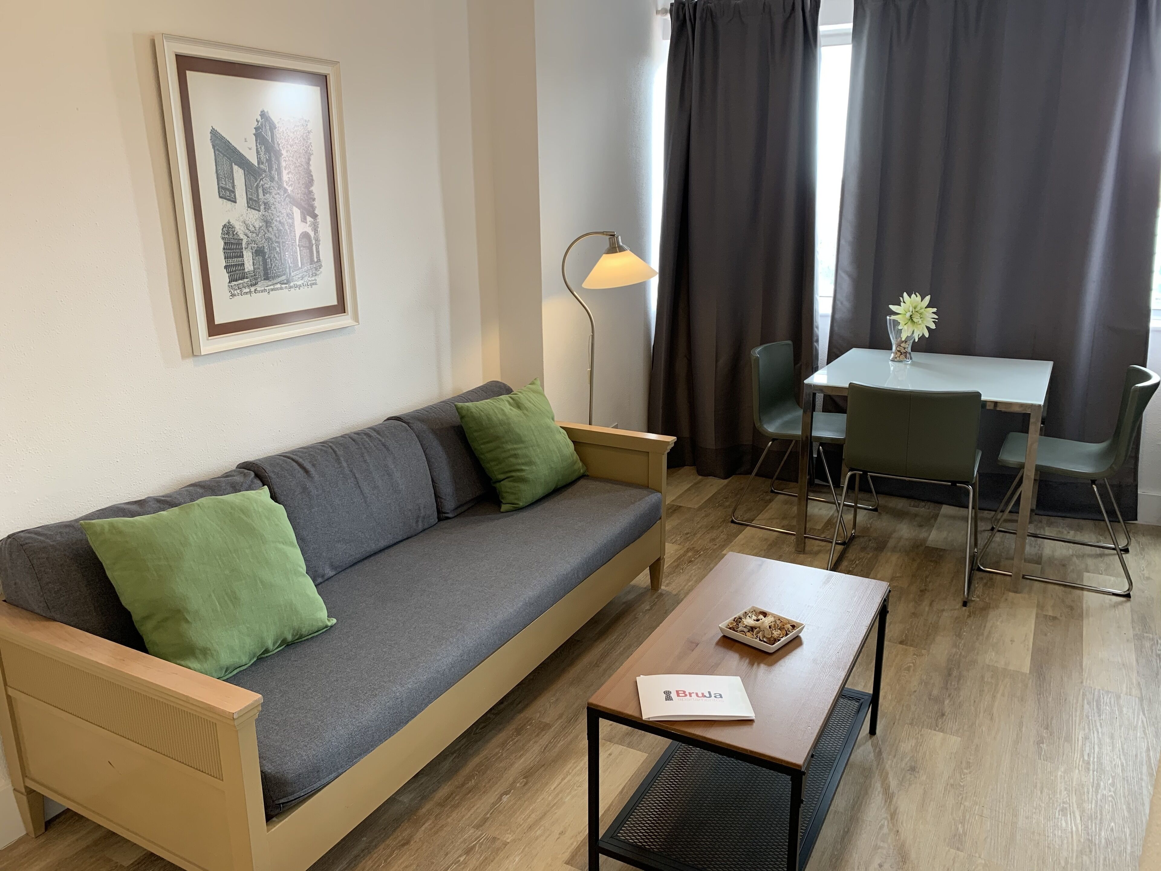 Ett modernt vardagsrum med en soffa, ett matbord och stolar. På väggen hänger en inramad tavla, det finns en golvlampa och ett fönster med gardiner.