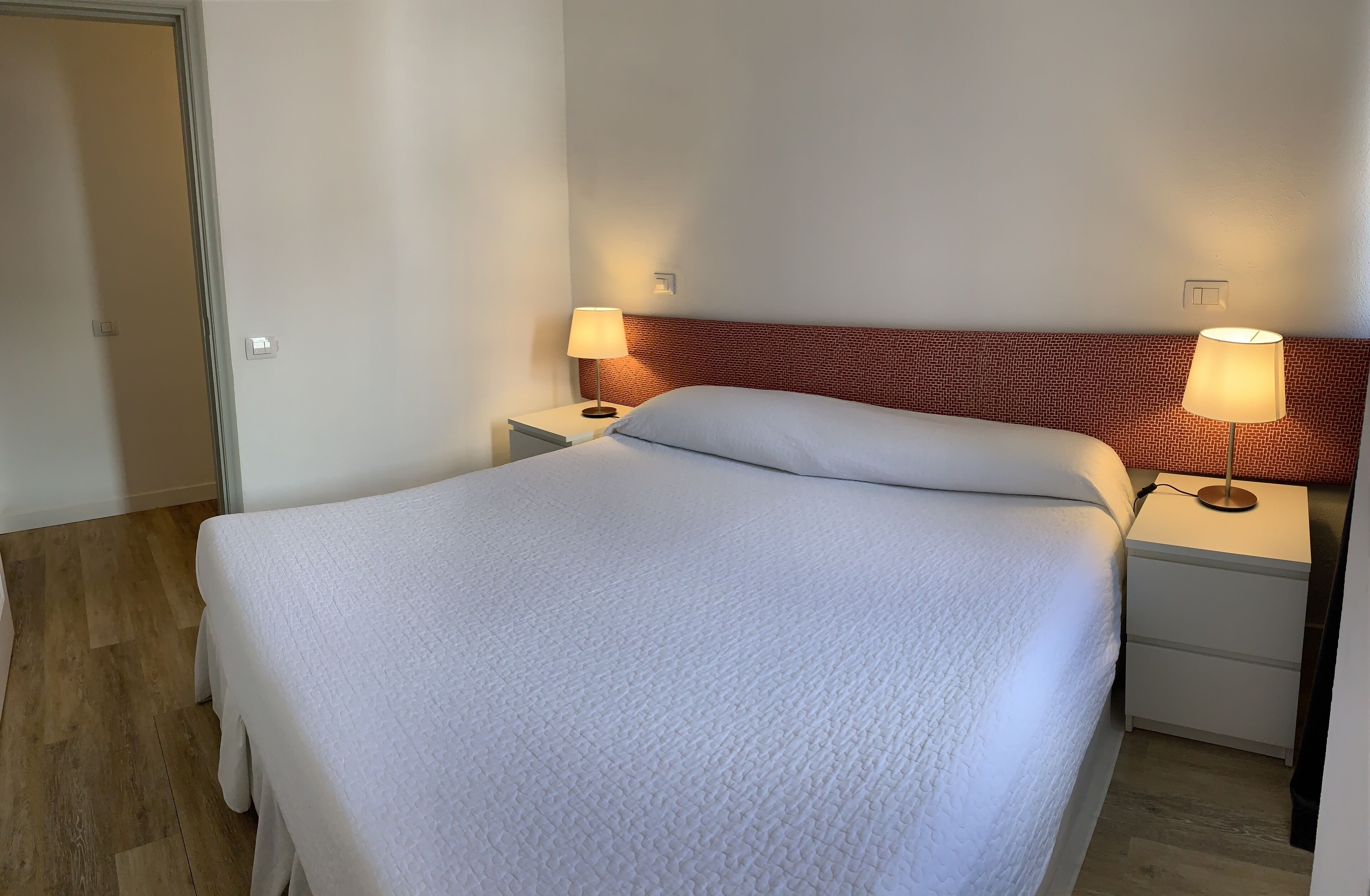 Una cama bien hecha con colcha blanca, dos mesitas de noche con lámparas y suelo de madera.