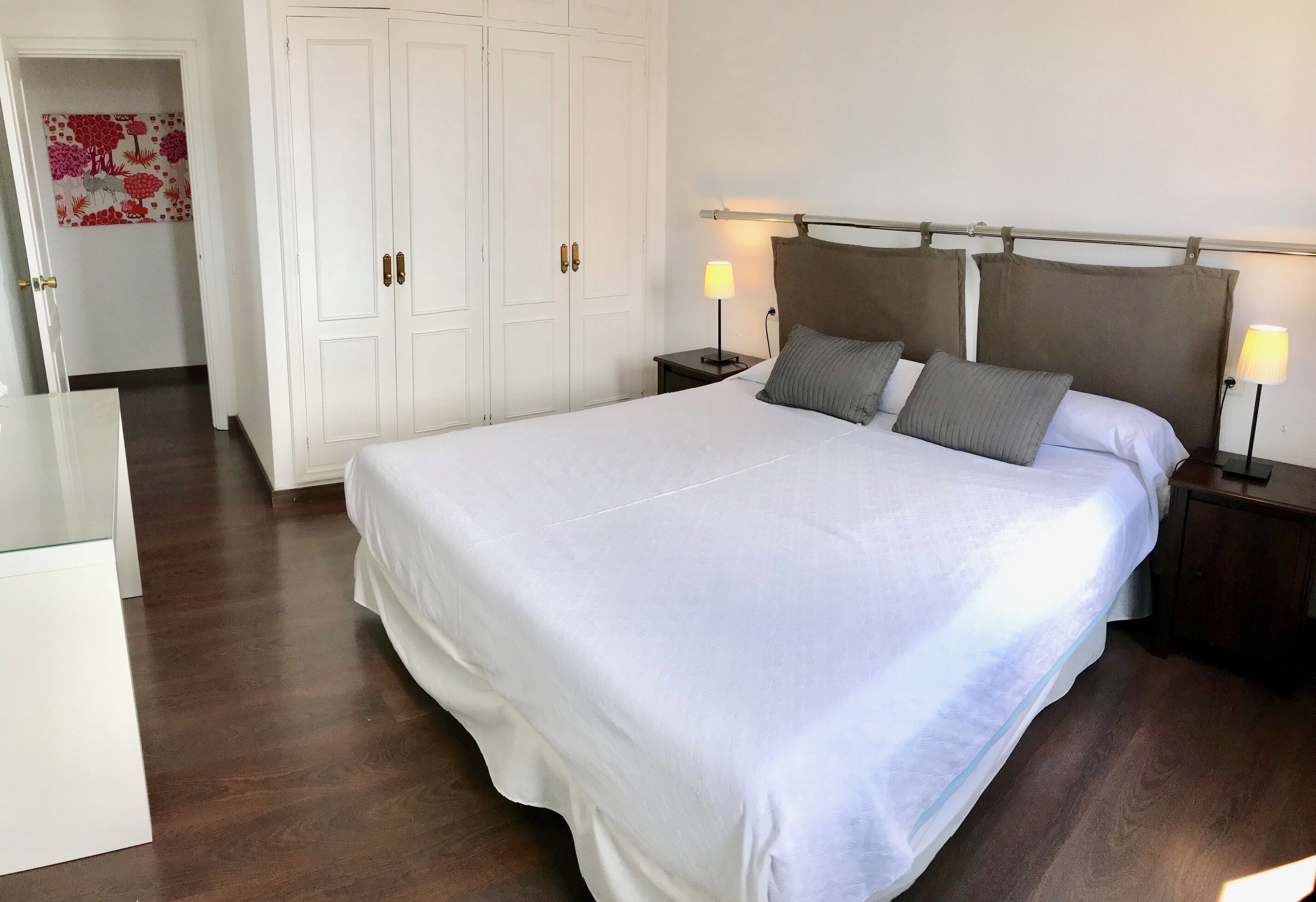 A neatly made bed with white linens, a headboard with a metal frame, two bedside lamps, and a large wardrobe with double doors.