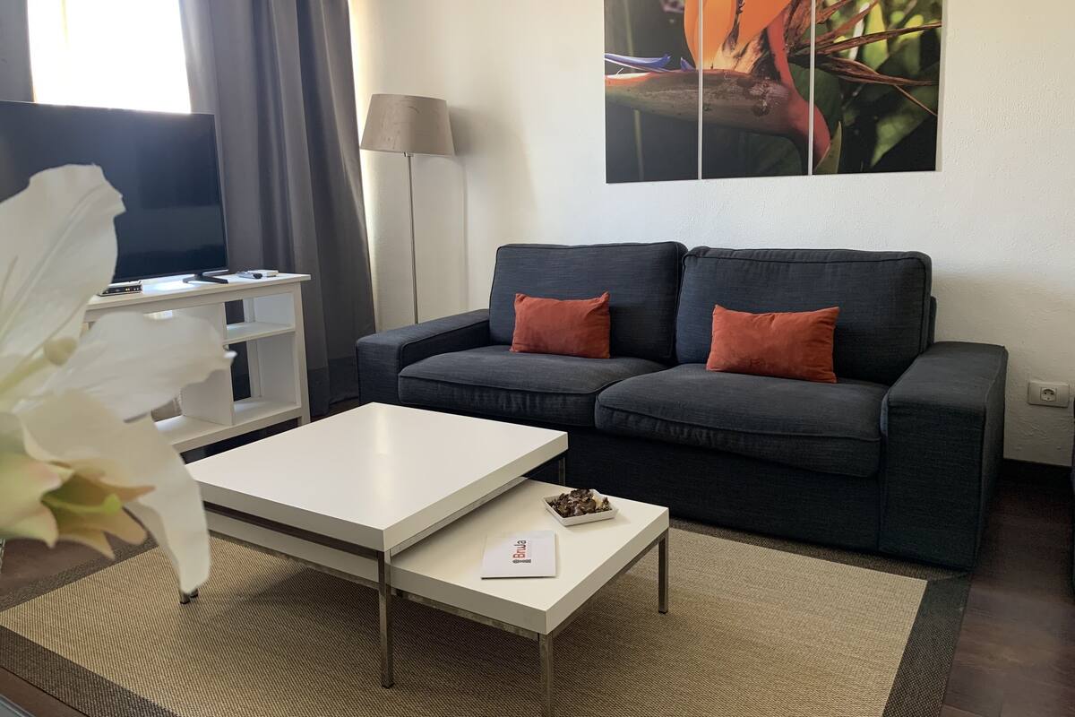 Ein Wohnzimmer mit einem dunkelgrauen Sofa, zwei orangenen Kissen, einem weißen Couchtisch und einer Stehlampe.