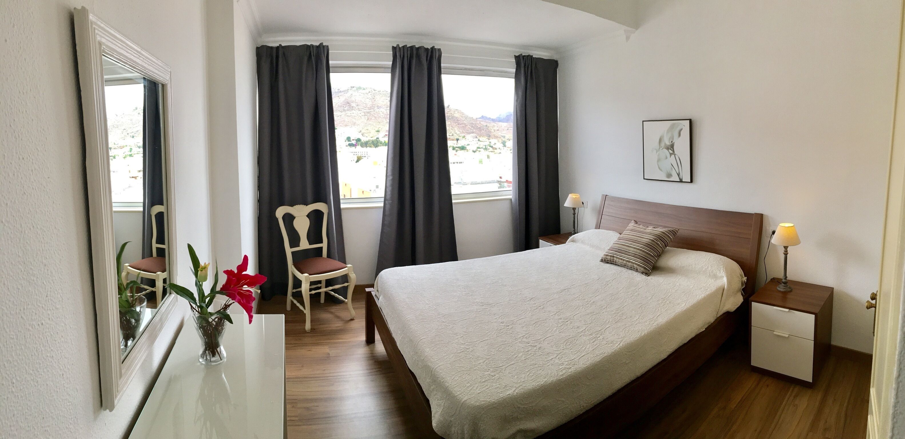 A bedroom with a large bed, a chair, a nightstand with a lamp, a bedside table, a mirror, and a window with curtains.