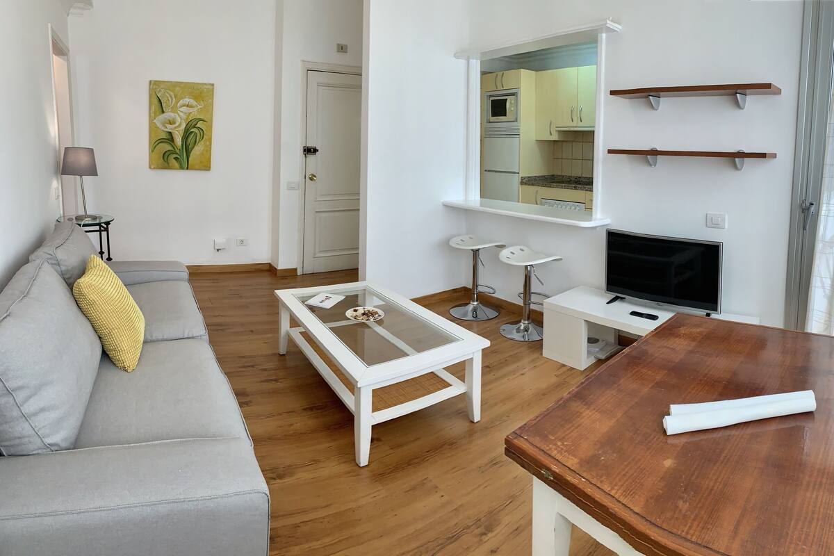 Ein modernes Wohnzimmer mit einem grauen Sofa, einem Couchtisch, einem Holzessttisch, einem Fernseher und einer Küche im Hintergrund.