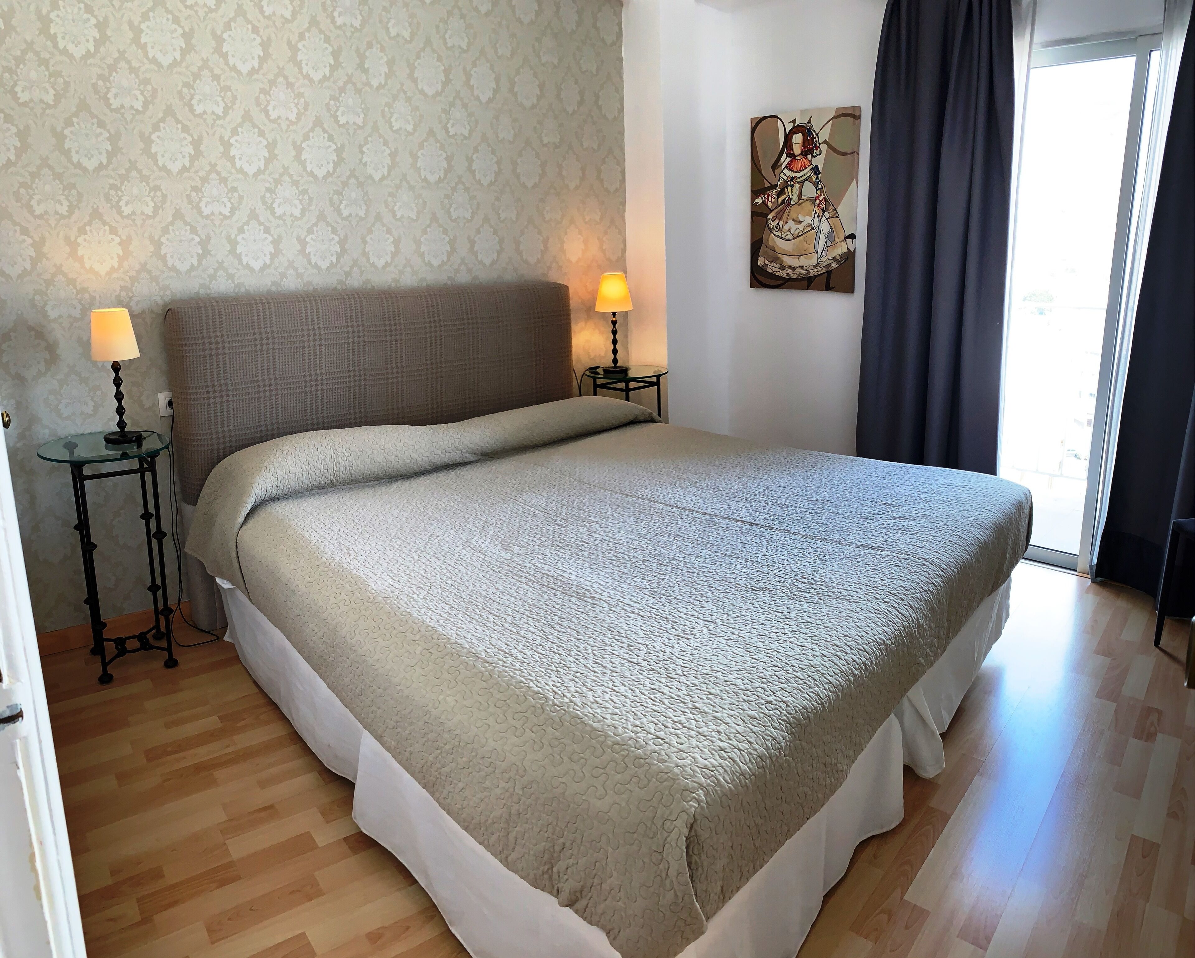 A bedroom with a bed, two bedside lamps, a patterned wallpaper, a painting on the wall, and a window with curtains.