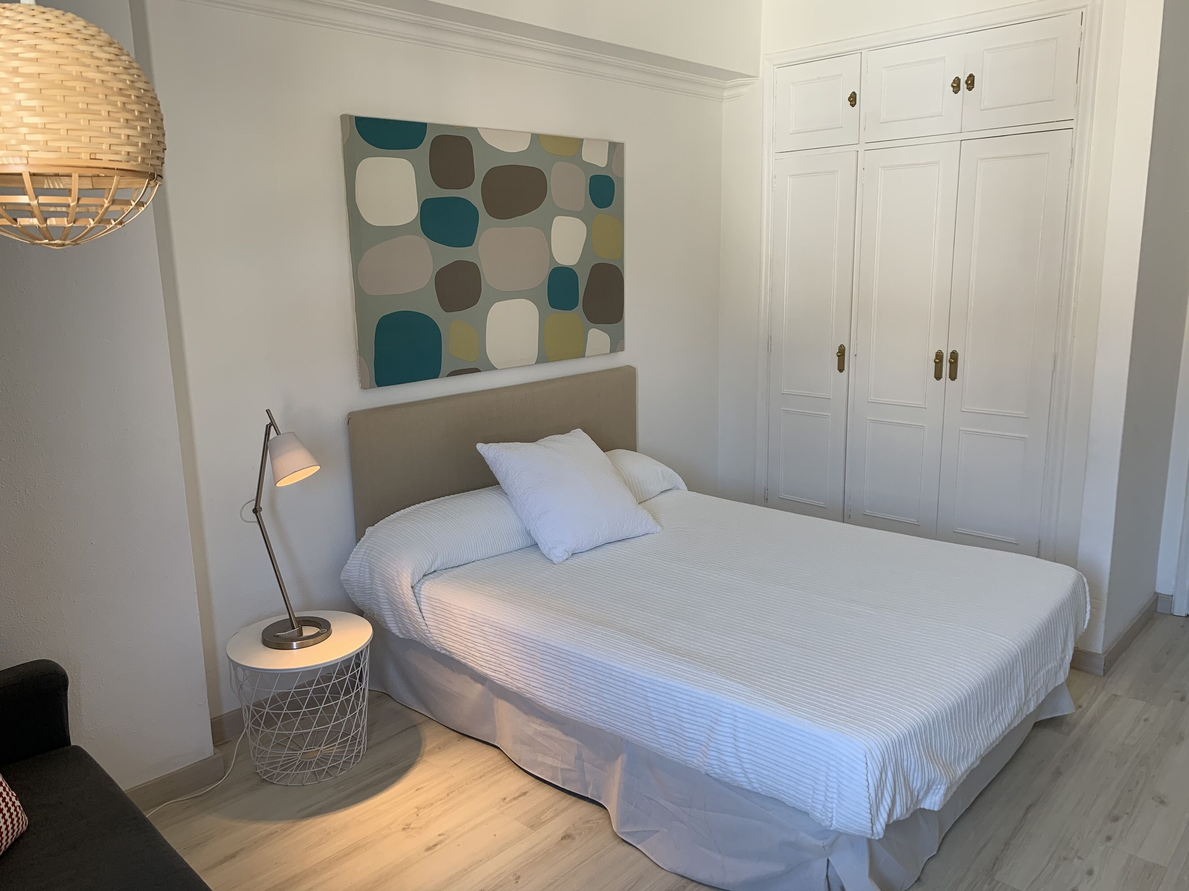 A bedroom with a bed, a bedside lamp, a round nightstand, a wall art piece, and a wardrobe.