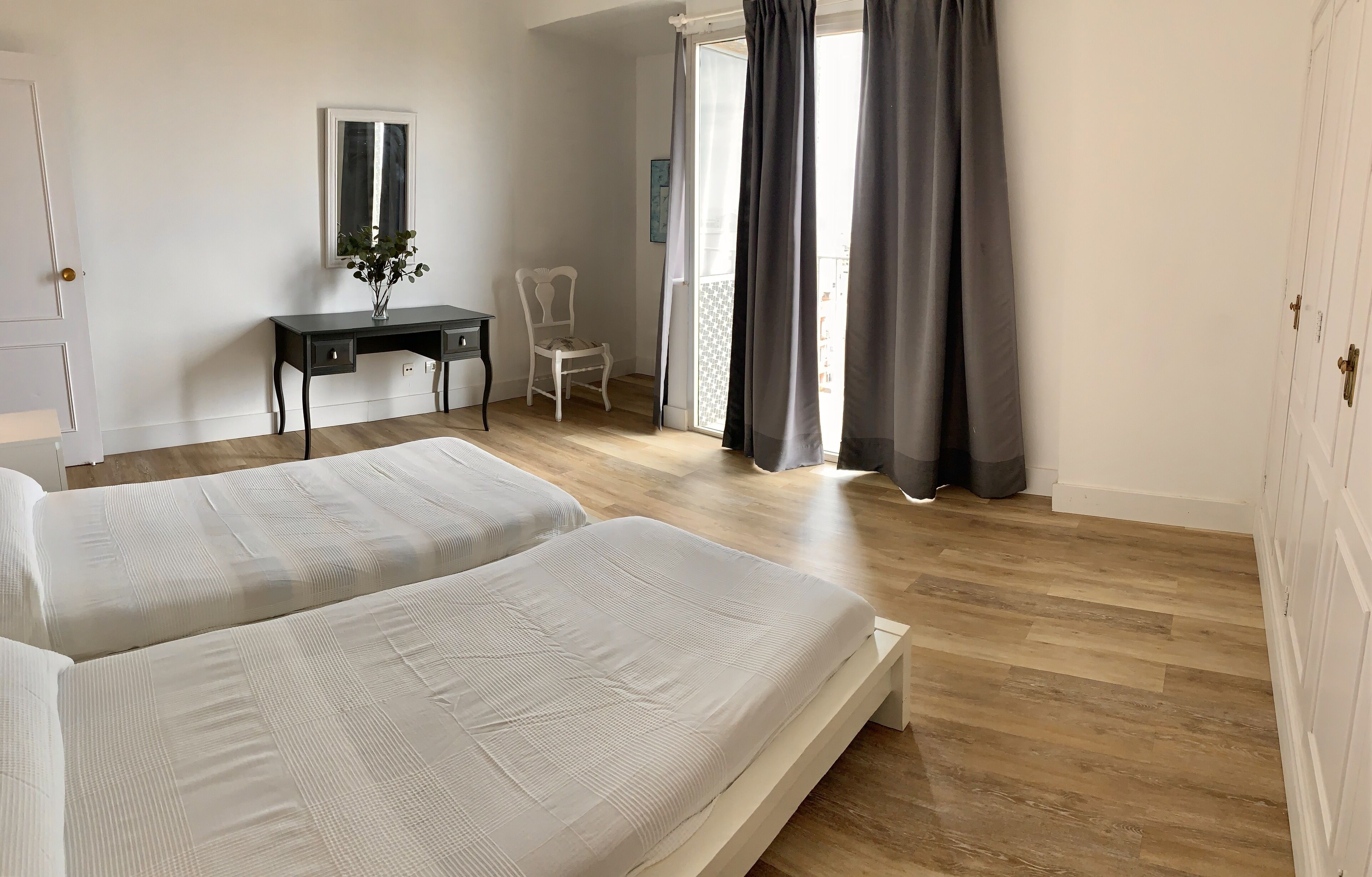 A bedroom with two beds, a desk, a chair, and a balcony with curtains.