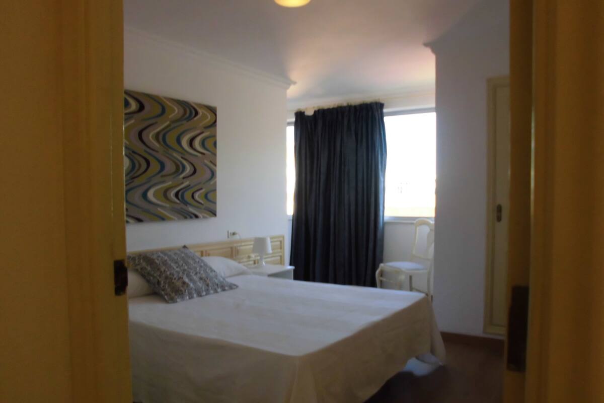 Ein Schlafzimmer mit Bett, Stuhl, Fenster mit Vorhängen und einer Wand mit einem farbenfrohen, abstrakten Gemälde.