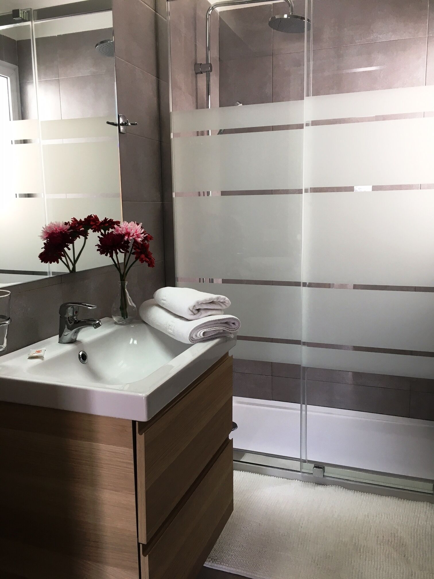 A modern bathroom with a glass shower enclosure, a white sink with a mirror, and a vase of flowers.