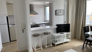Eine kompakte Küche mit eingebautem Herd, einer Mikrowelle und einer Dunstabzugshaube. Zwei Barhocker stehen an einem weißen Tisch mit einer dekorativen Mittelstück. Ein Flachbildfernseher ist über dem Tisch an der Wand angebracht.