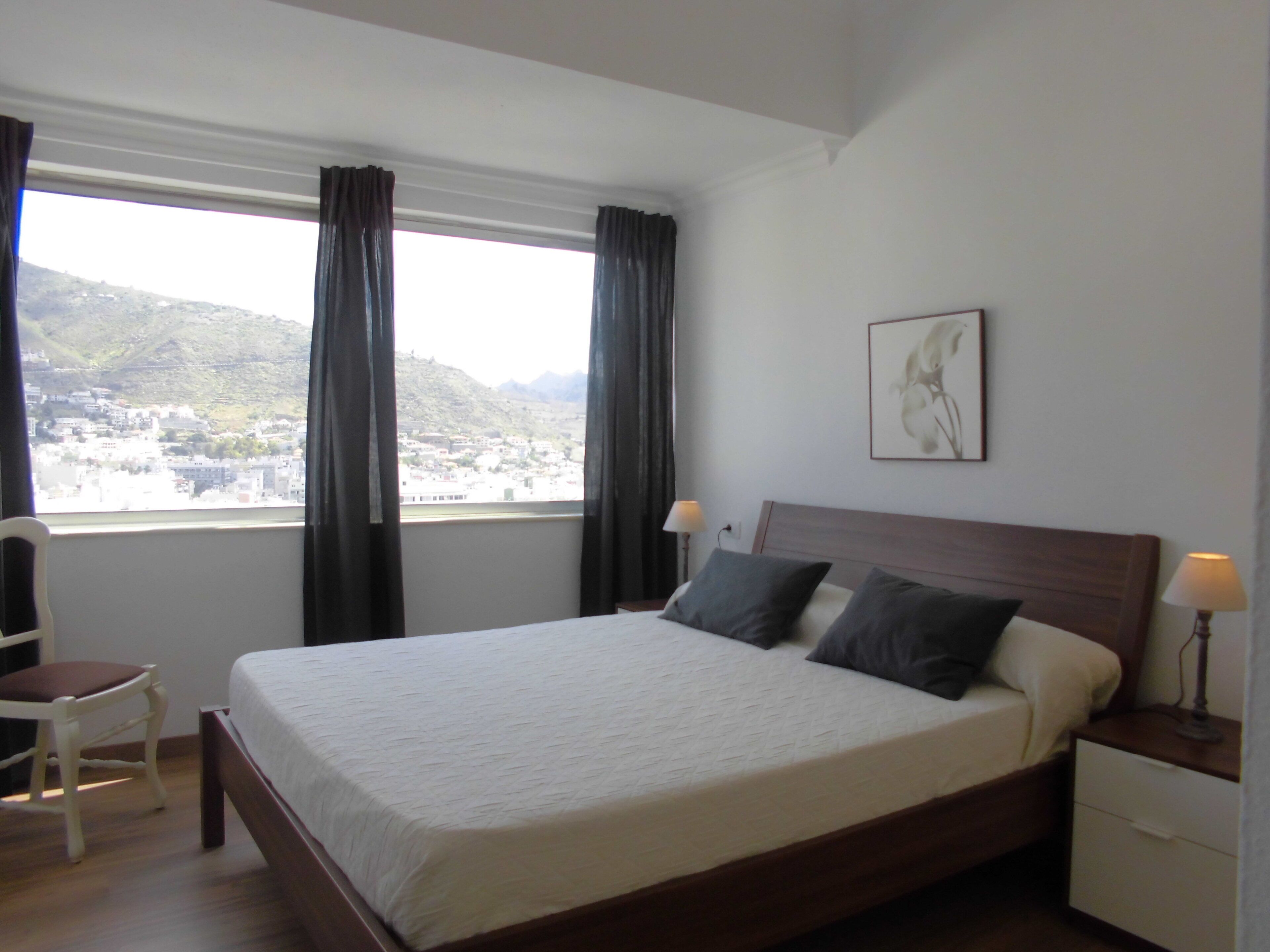 Een netjes opgemaakt bed met een houten hoofdbord, een nachtkastje met een lamp en een stoel. Een raam met gordijnen biedt uitzicht op een stad en heuvels.