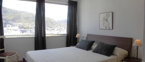 A neatly made bed with a wooden headboard, a bedside table with a lamp, and a chair. A window with curtains provides a view of a town and hills.