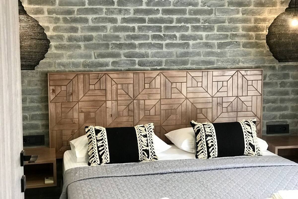 Habitación de hotel con pared de ladrillo, cabecera de madera con diseño geométrico, cama con almohadas blancas y negras, y colcha acolchada gris.