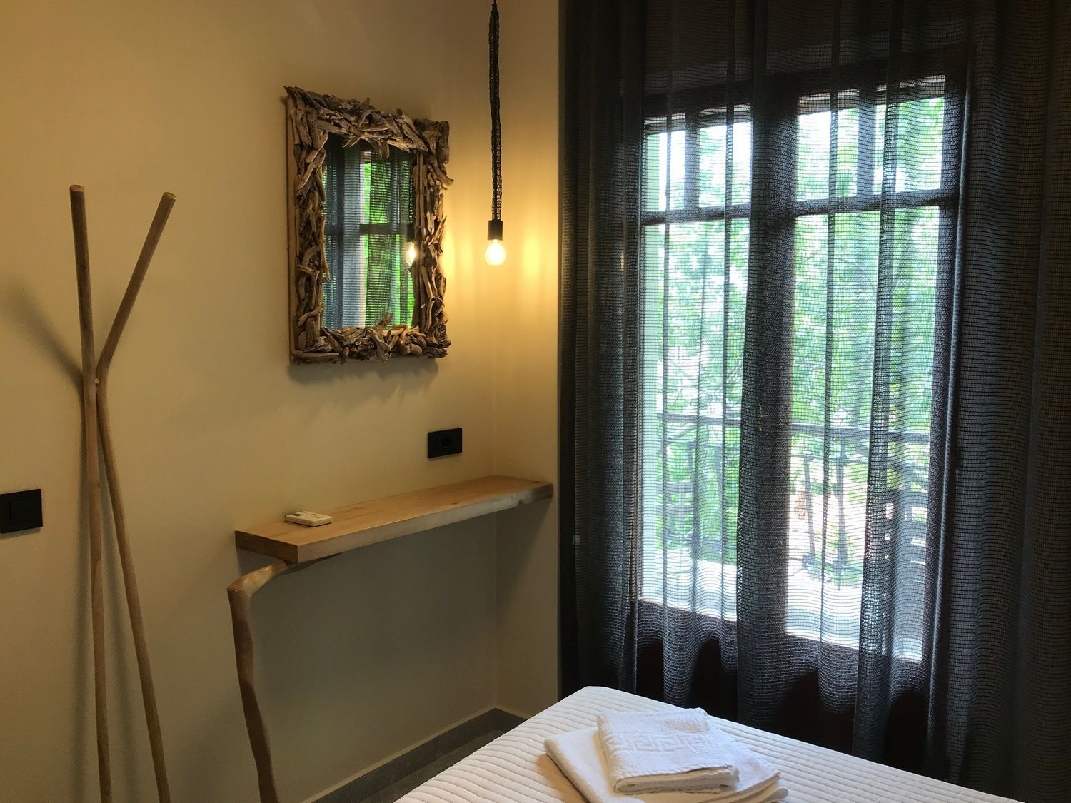 Hotellihuone, jossa on sänky, yöpöytä, peili ja ikkuna, jossa on läpikuultavat verhot.