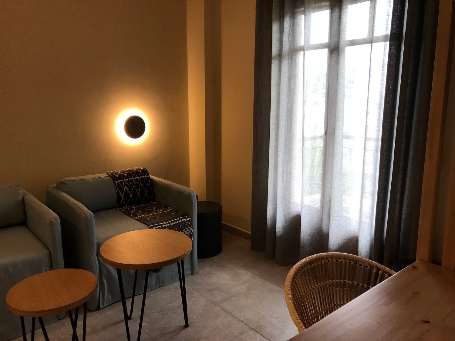 Una sala acogedora con un sofá, dos mesitas, un sillón de mimbre y una lámpara de pared redonda.