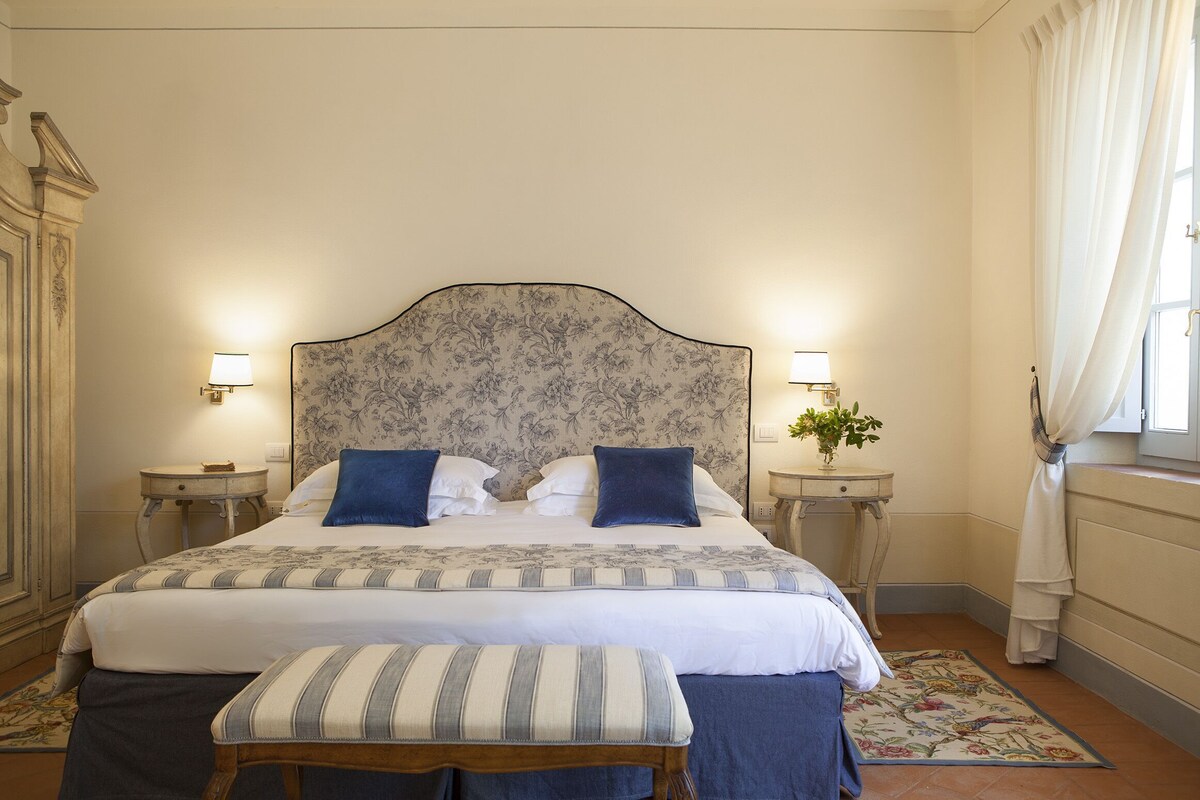 A bedroom with a large bed, two bedside tables with lamps, a bench, and a floral-patterned rug.