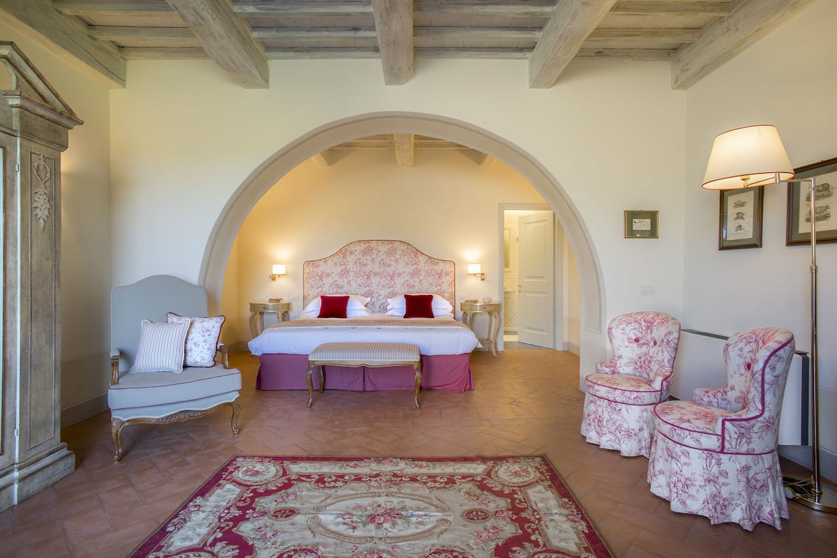 A bedroom with a large bed, two chairs, a bench, and a floor lamp.