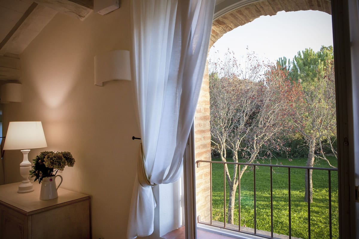 A room with a view of a garden, a lamp on a bedside table, and a window with curtains.