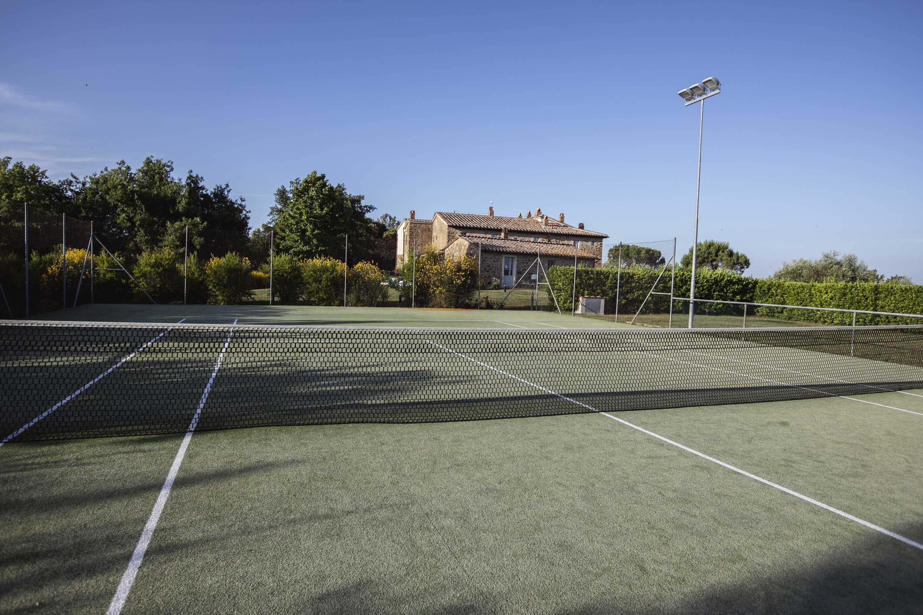An outdoor tennis court with a net, surrounded by greenery and a building in the background.