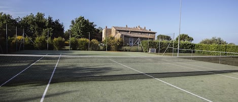 An outdoor tennis court with a net, surrounded by greenery and a building in the background.