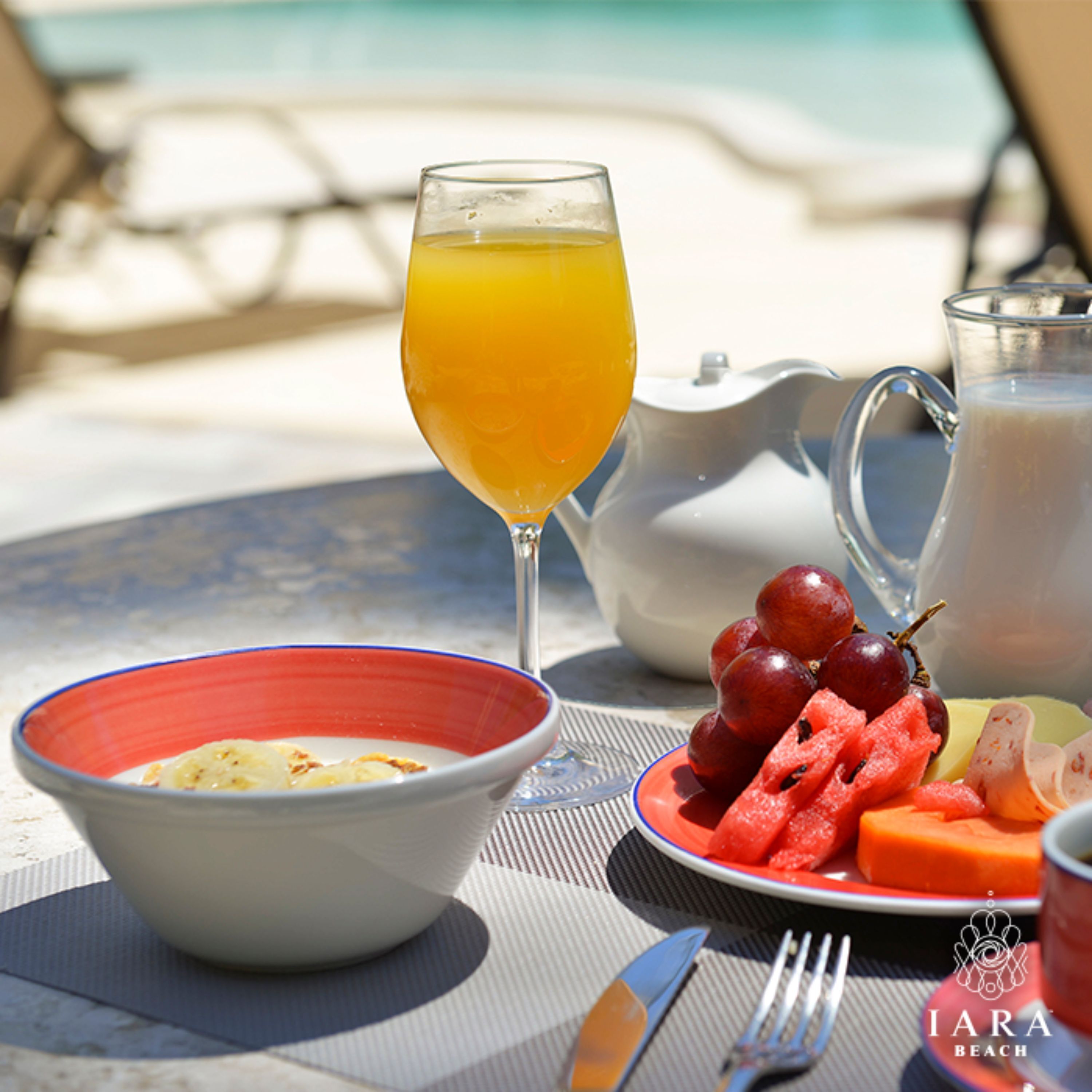 A breakfast table with a glass of orange juice, a teapot, a pitcher, a plate with grapes and watermelon, and a bowl of cereal.