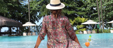 A person sitting by a pool with a tropical setting, including palm trees and umbrellas.