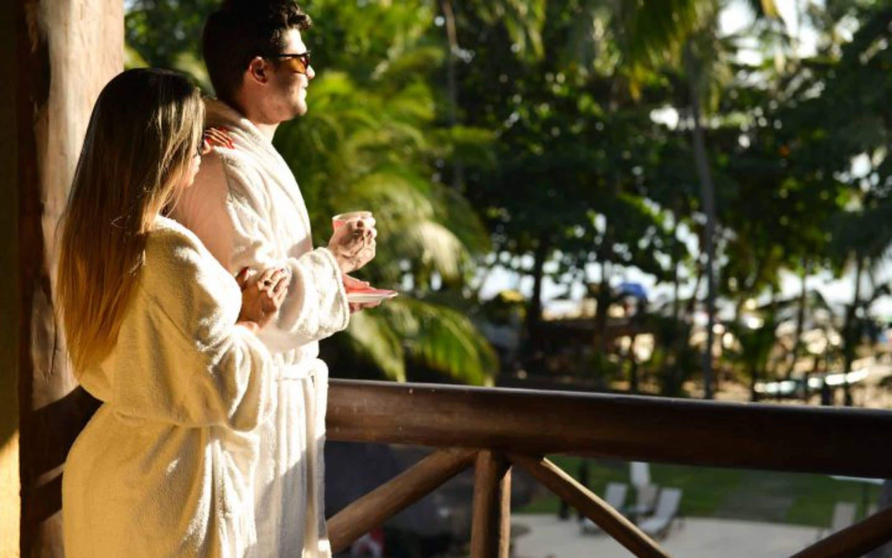 A couple standing on a balcony with a view of palm trees and a pool.