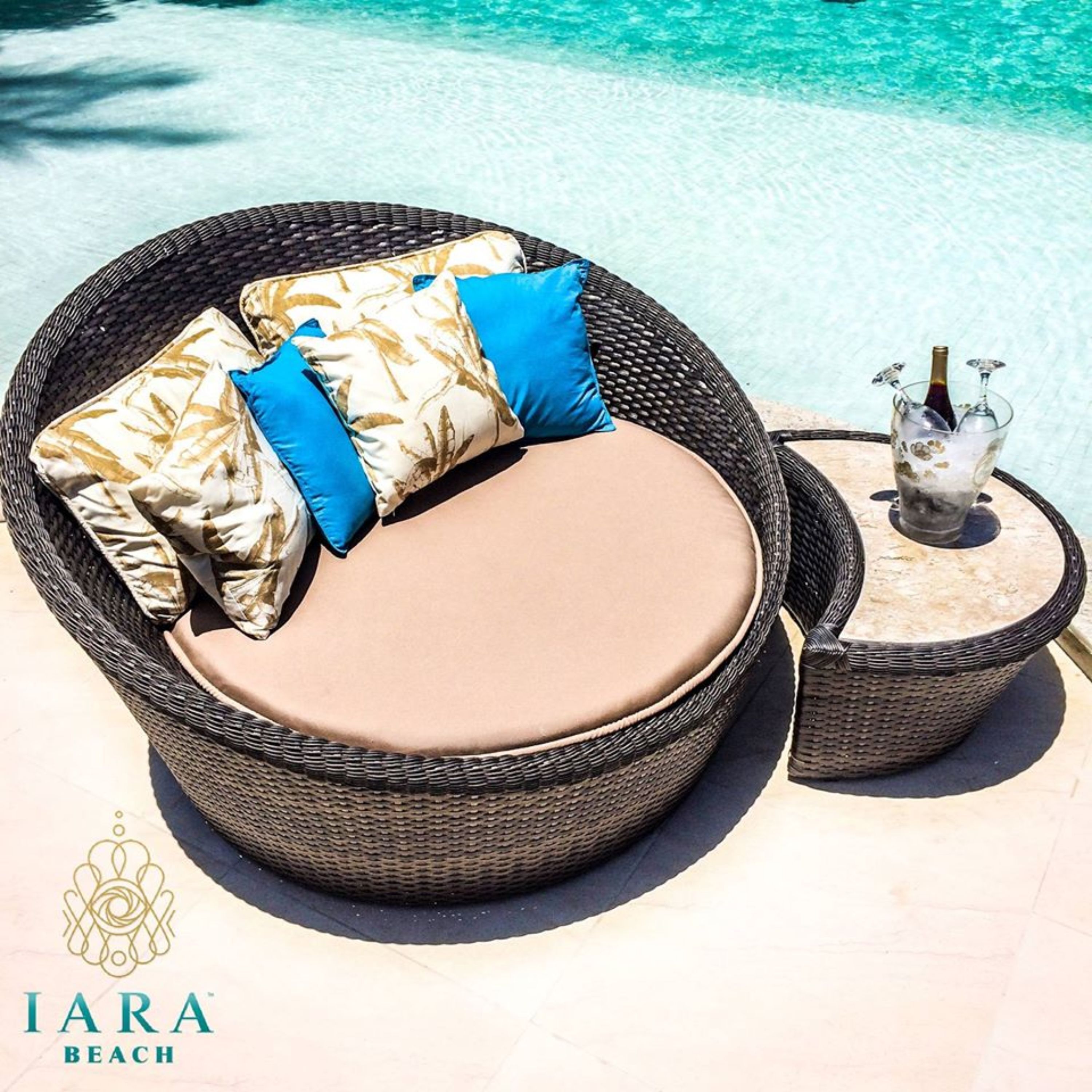 A wicker outdoor seating area with cushions, a small table with a bottle and glasses, and a clear view of the pool.