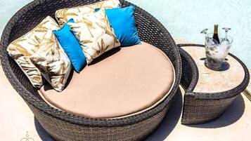 A wicker outdoor seating area with cushions, a small table with a bottle and glasses, and a clear view of the pool.