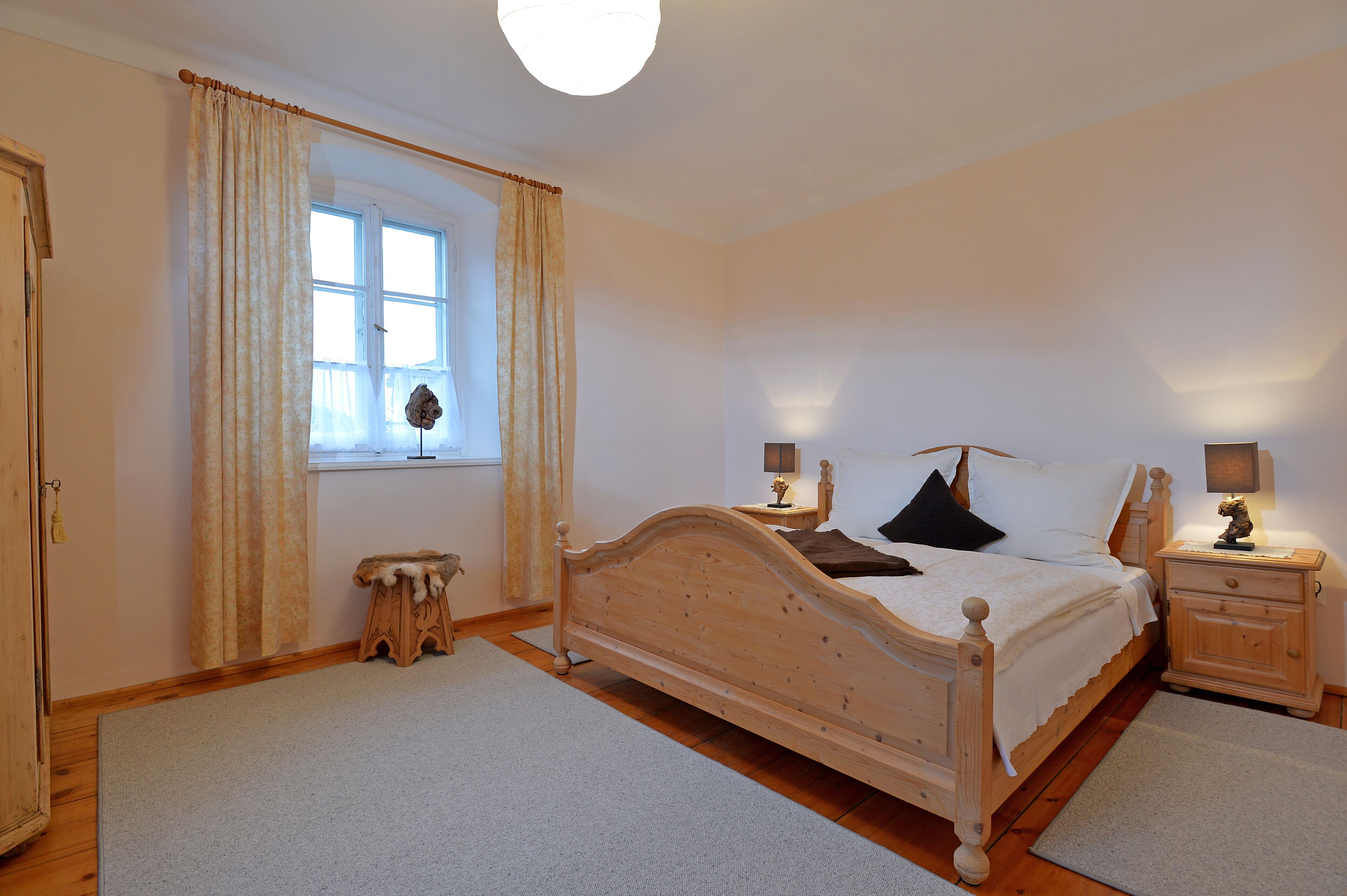 A bedroom with a wooden bed, two bedside tables with lamps, a window with curtains, and a small stool.