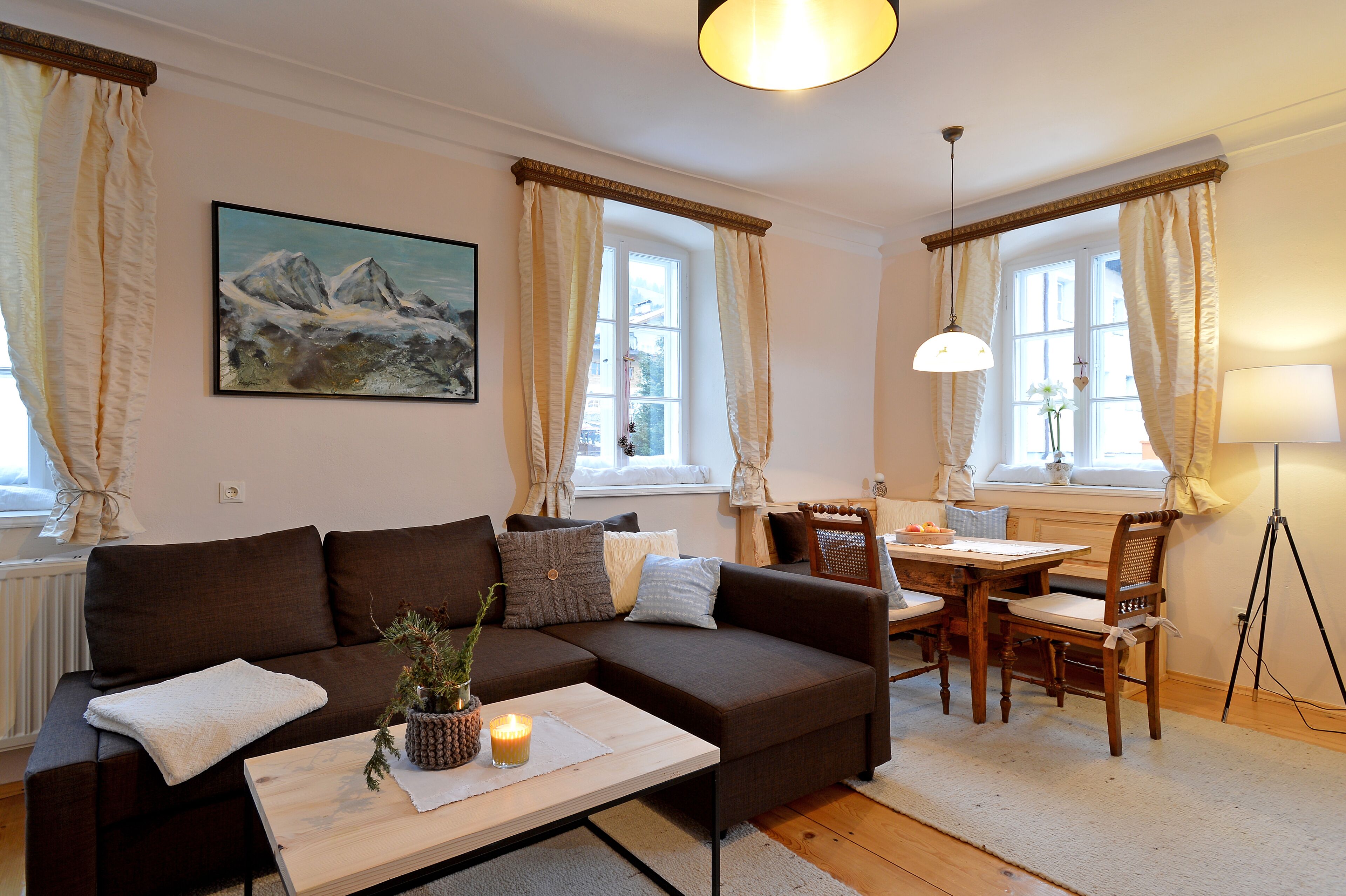 Ein gemütliches Wohnzimmer mit einem braunen Sofa, einem Esstisch mit Stühlen, einer Lampe und einem Bergpanorama an der Wand.