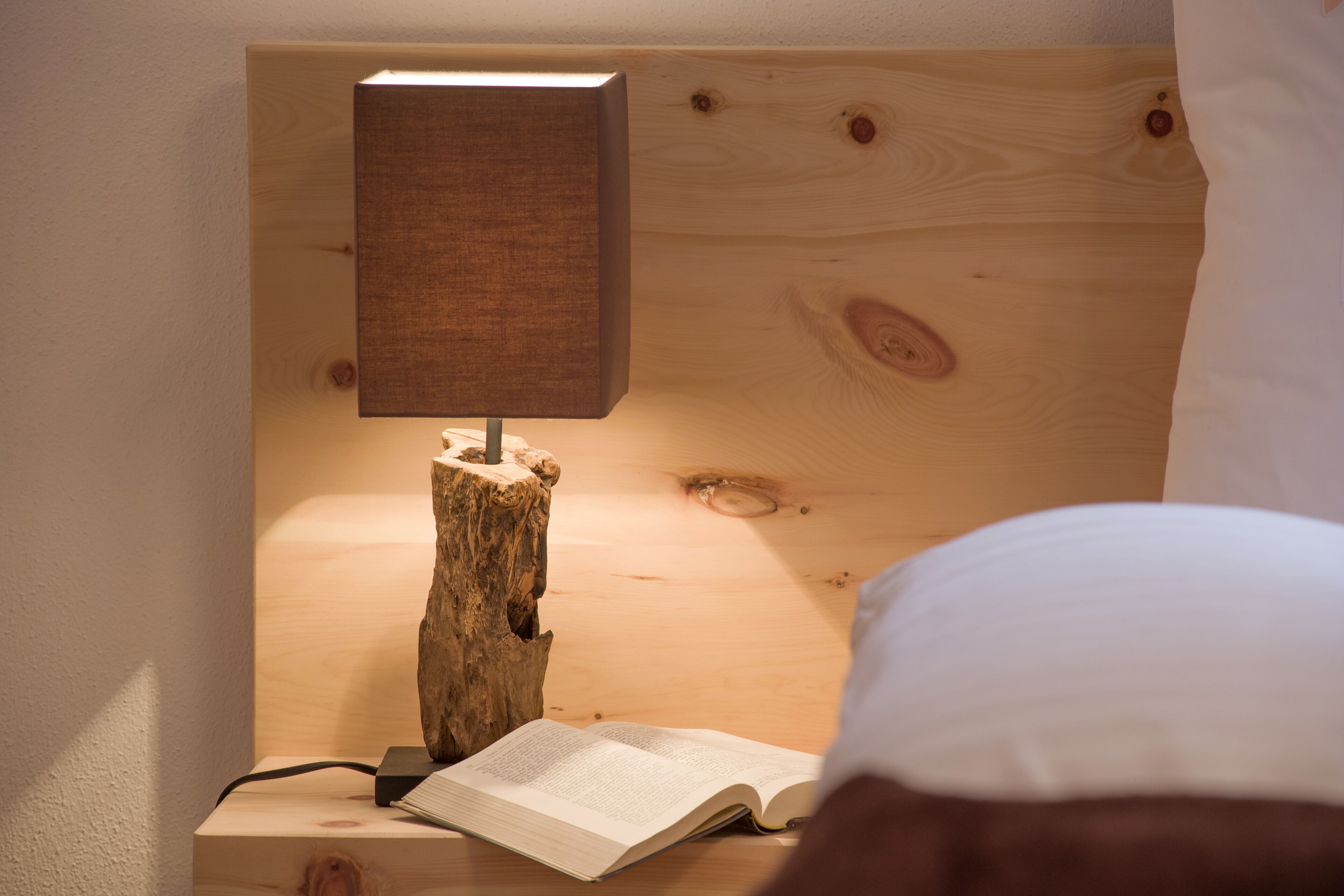 A wooden bedside table with a lamp, an open book, and a white pillow.