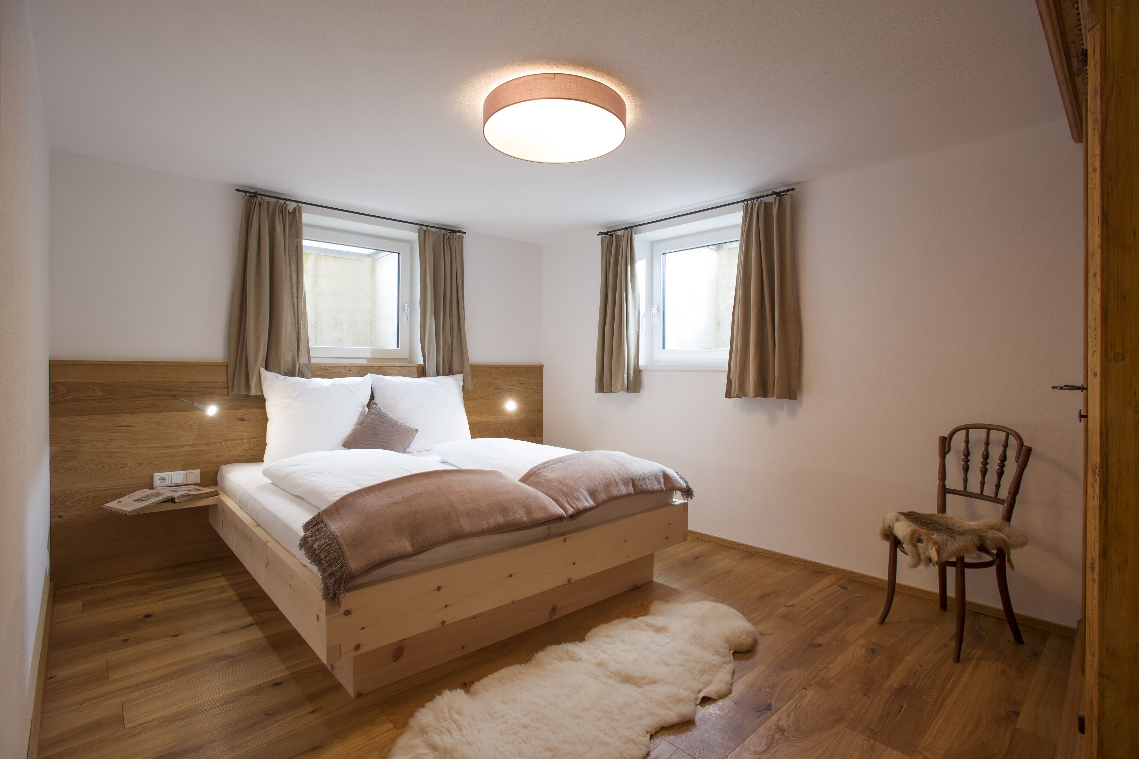 A bedroom with a wooden bed, a small chair, a fur rug, and two windows with curtains.