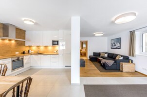 Una cocina moderna con armarios blancos, encimera de madera y electrodomésticos integrados. Cuenta con una zona de comedor con mesa y sillas de madera. La sala de estar presenta un sofá de color oscuro y un aparador de madera.