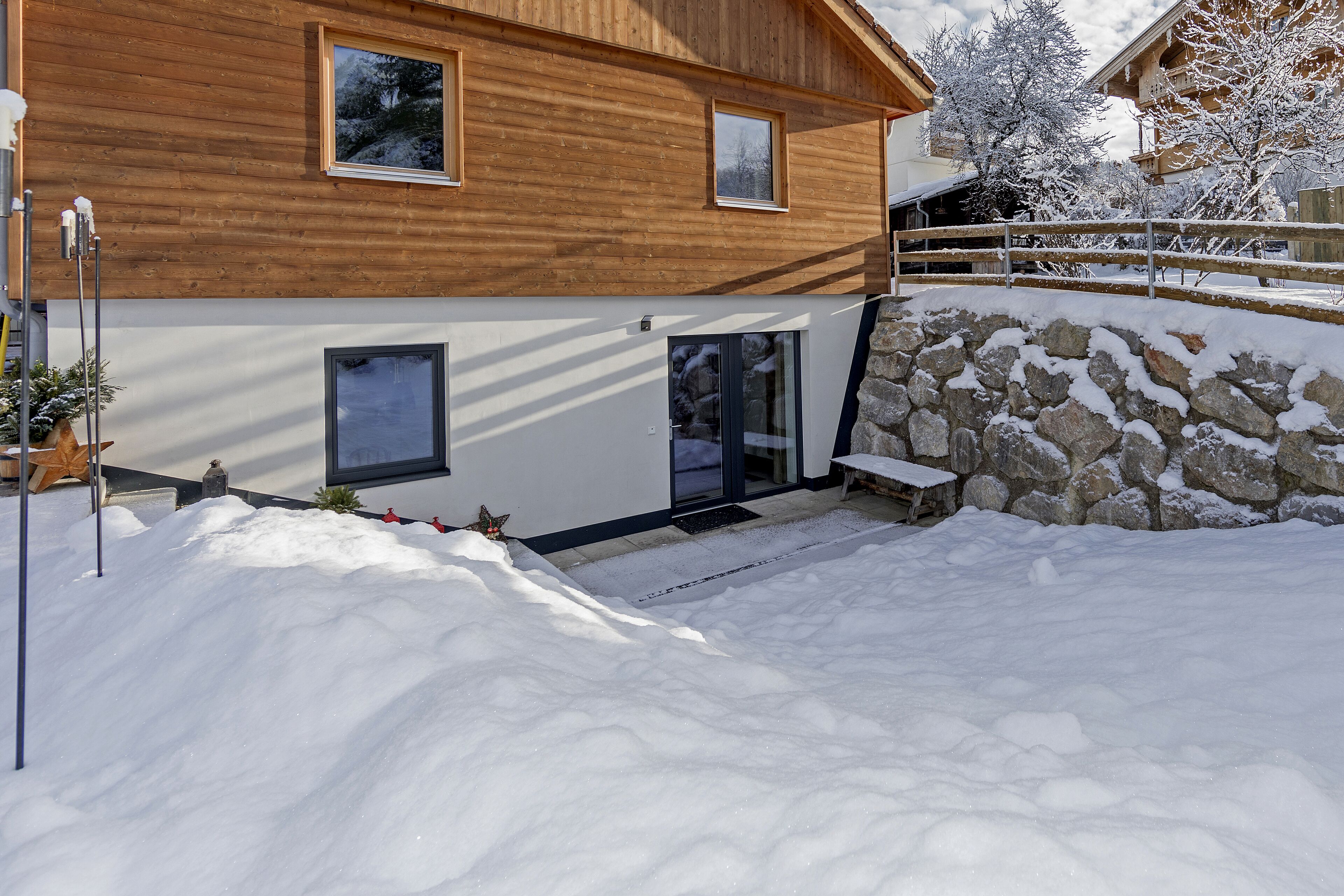 Una casa de madera con un muro de piedra y el suelo cubierto de nieve.