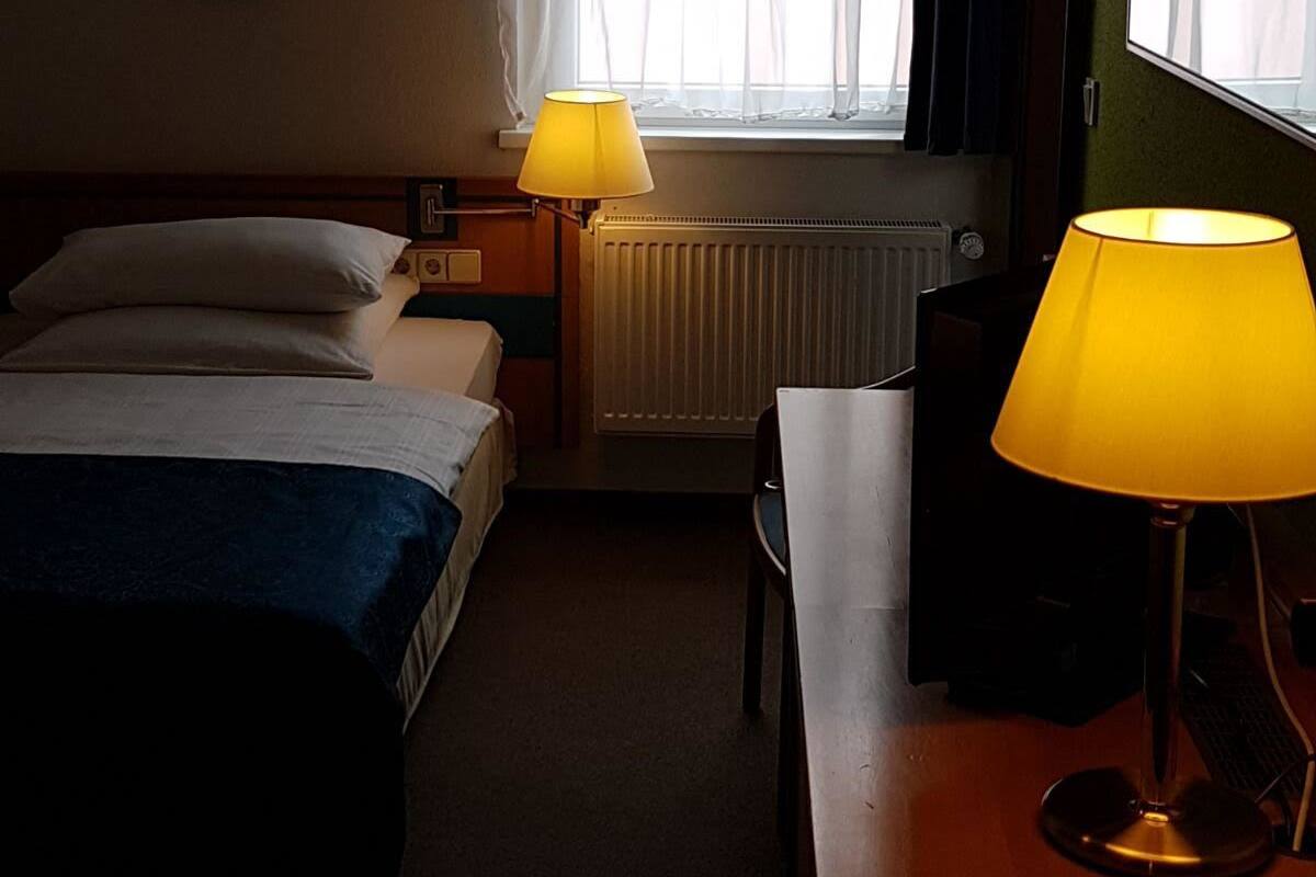 A hotel room with two beds, a desk with a lamp, a radiator, and a window with sheer curtains.