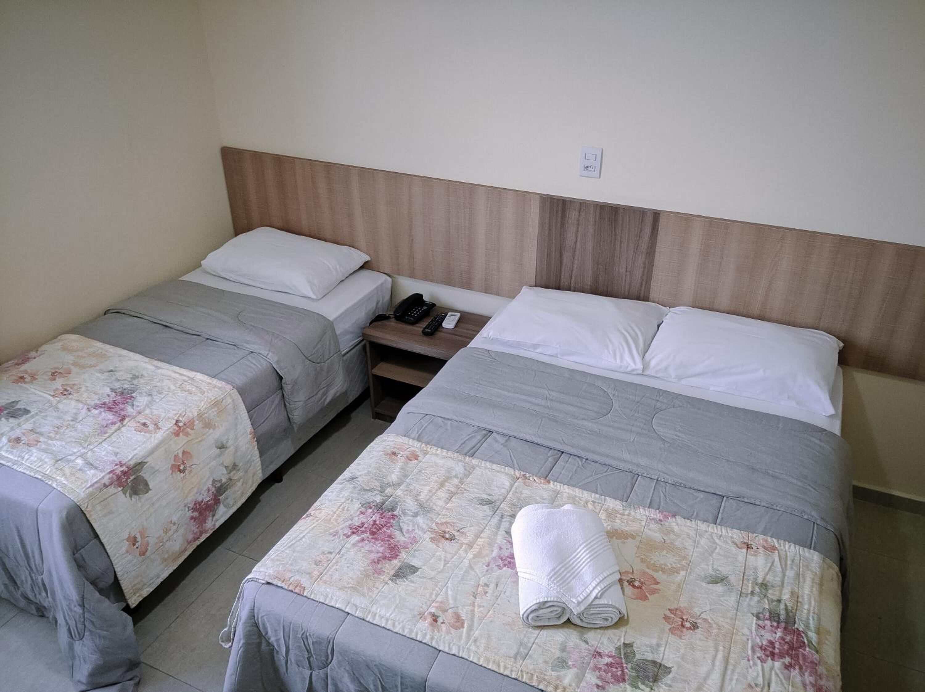 A hotel room with two single beds, a wooden headboard, a bedside table with a phone and remote control, and a folded towel on the bed.