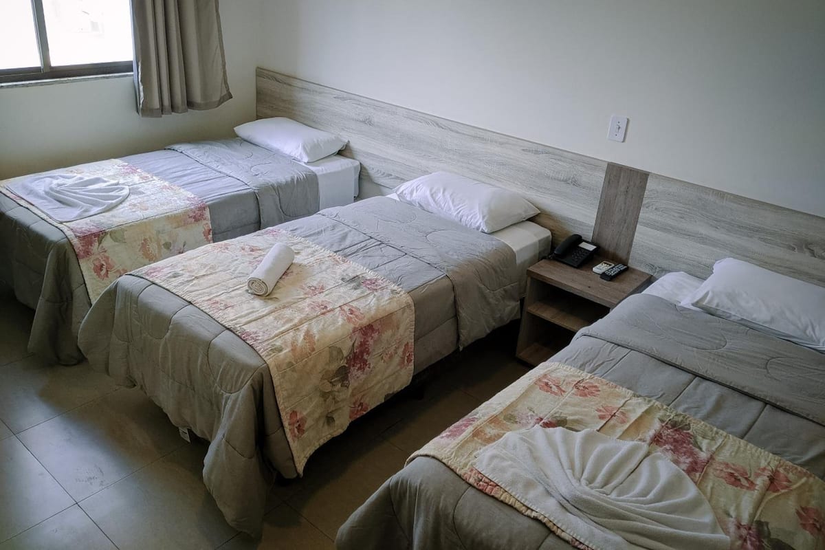 Habitación de hotel con tres camas individuales, cada una con cubrecama floral y almohadas blancas. Se observa una mesita de noche de madera con un teléfono y un control remoto.