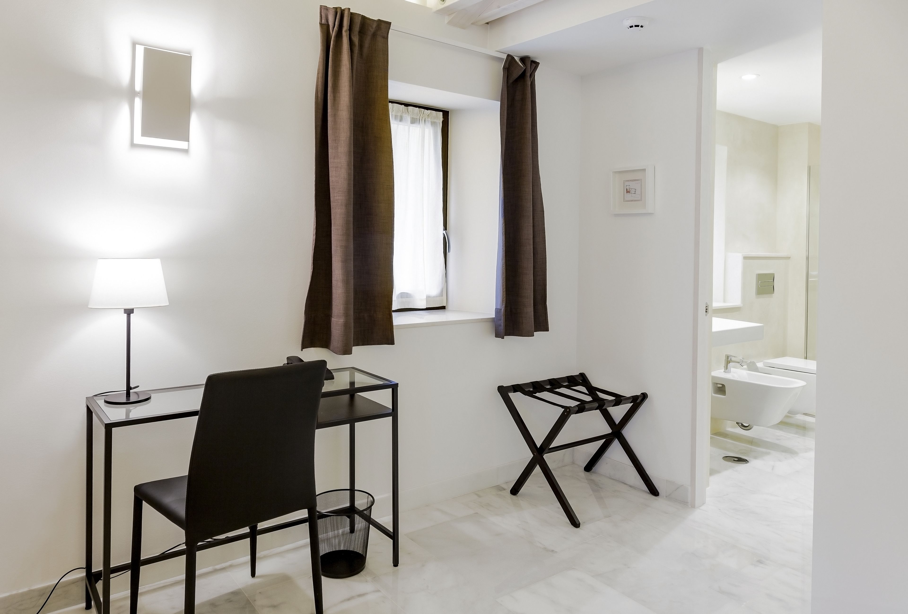 Une petite pièce moderne, équipée d’un bureau, d’une chaise et d’une petite table. On y voit une fenêtre avec des rideaux, une lampe, et, au fond, une salle de bain.