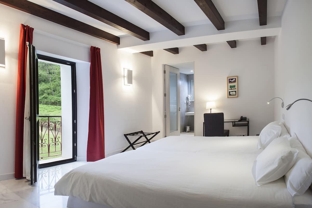 A hotel room with a large bed, a desk, a chair, a bathroom, and a balcony with red curtains.