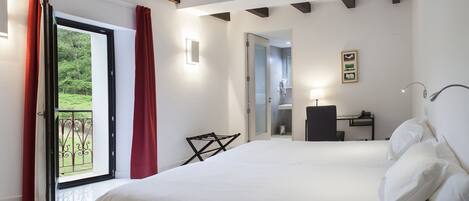 A hotel room with a large bed, a desk, a chair, a bathroom, and a balcony with red curtains.