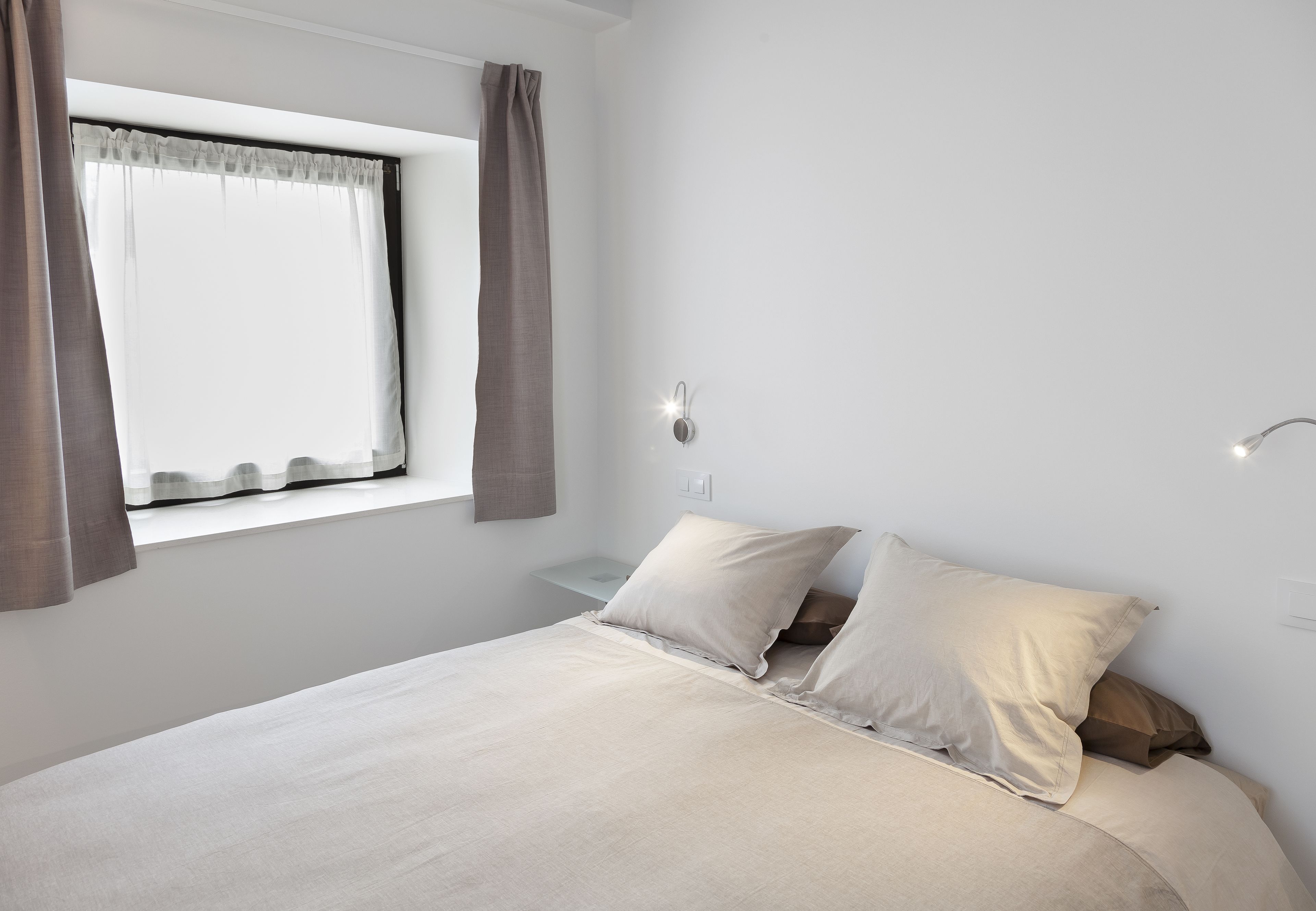 Um quarto com uma cama grande, uma janela com cortinas, uma mesinha de cabeceira e uma luminária fixada na parede.