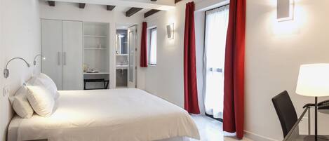 A modern bedroom with a large bed, a wardrobe, a desk with a lamp, and a view of the outside through a window with red curtains.