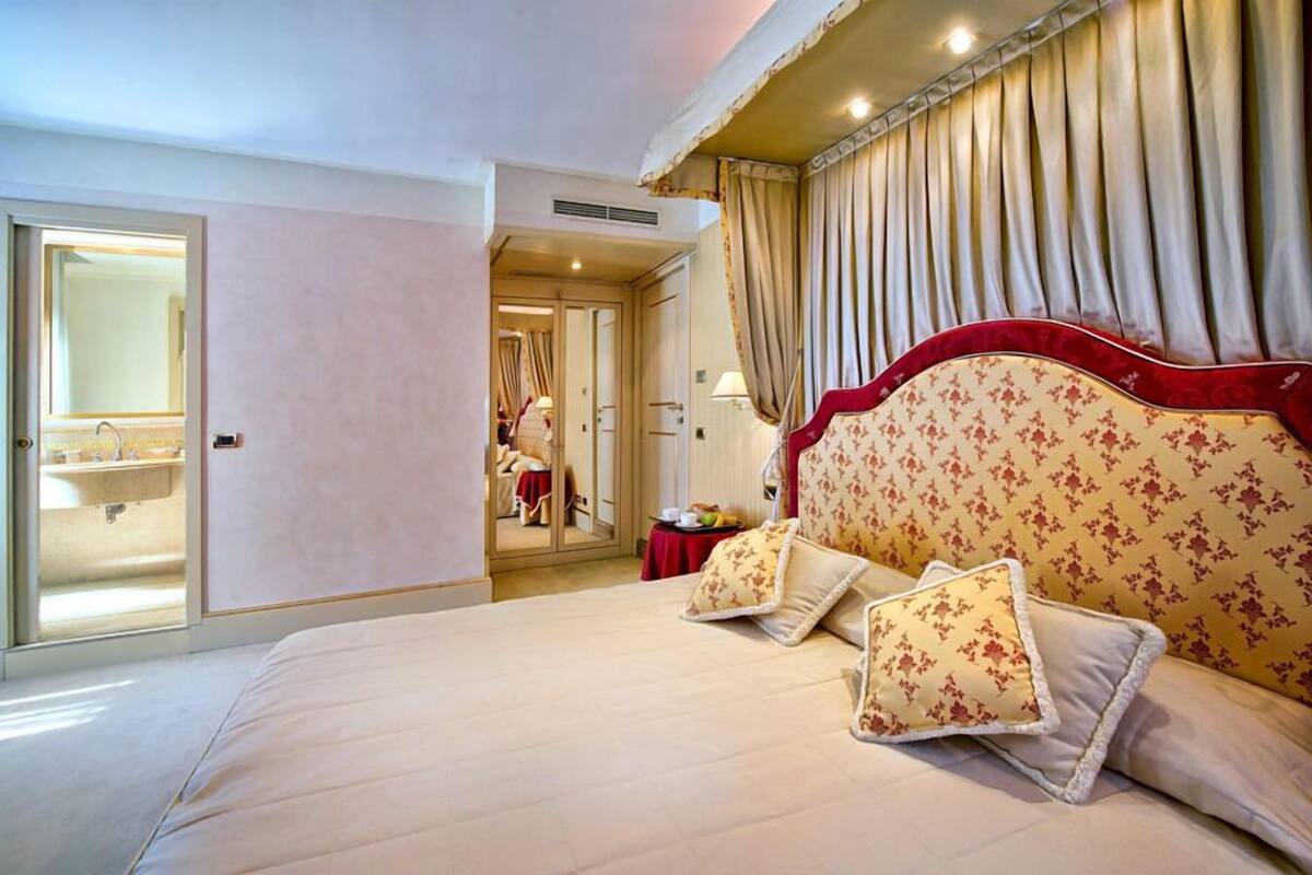 Une chambre d’hôtel avec un grand lit, un miroir, une coiffeuse avec un lavabo et une porte donnant sur une autre pièce.
