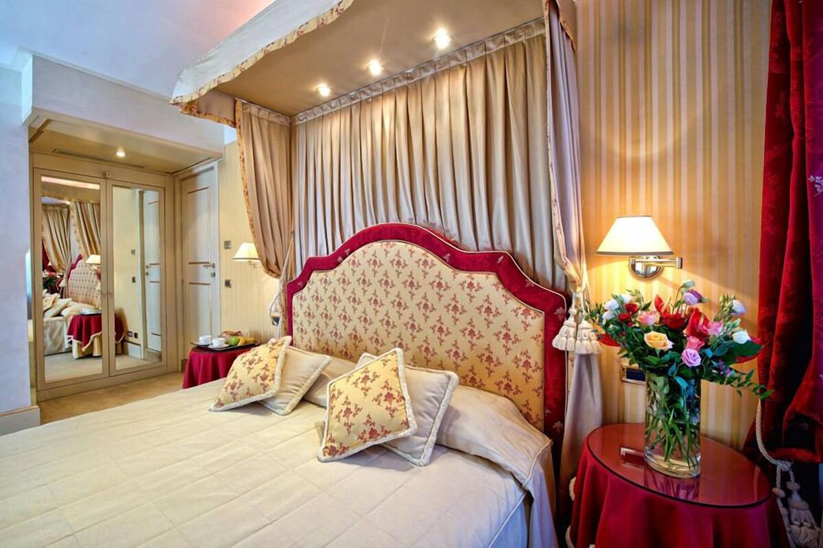 Une chambre d’hôtel avec un grand lit, un miroir, une table de chevet avec une lampe et un vase de fleurs.