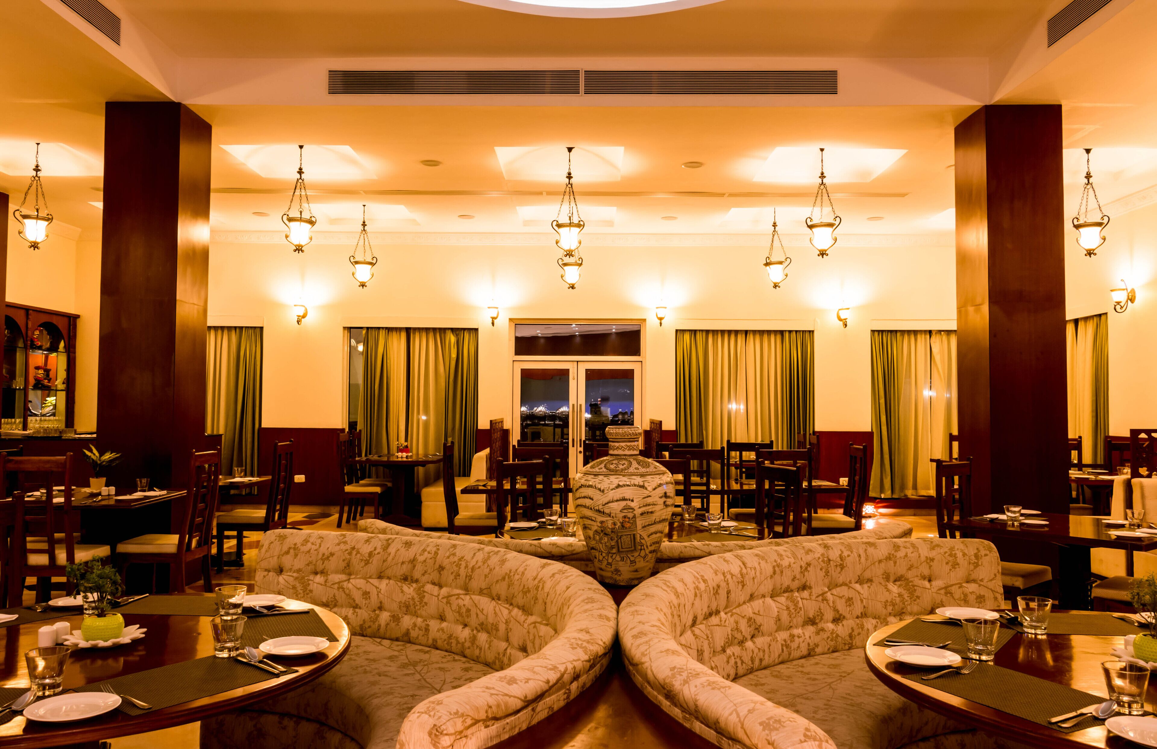 A dining area with multiple tables and chairs, a large vase centerpiece, and hanging chandeliers.