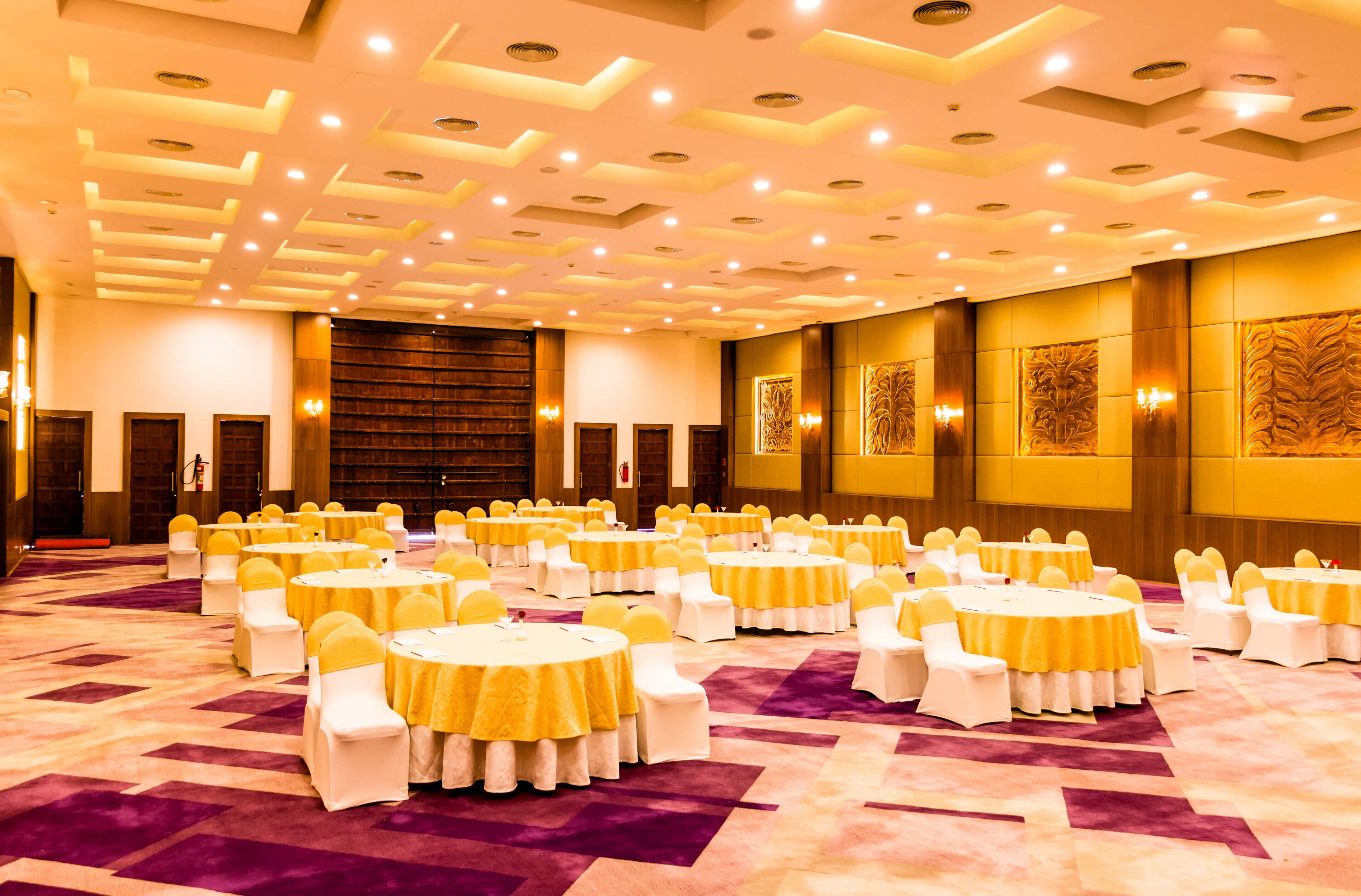 A large conference room with multiple round tables, each with white chairs and yellow sashes. The room features a patterned carpet and a ceiling with recessed lighting.